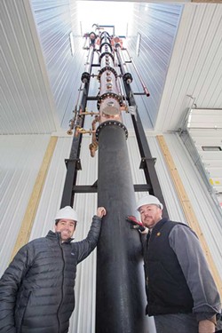 Jeremy Kidde and Jason Grizzanti stand in front of the newly installed Vendome continuous column still, about 50 feet tall and 18 feet in diameter, at Black Dirt Distillery in Pine Island.