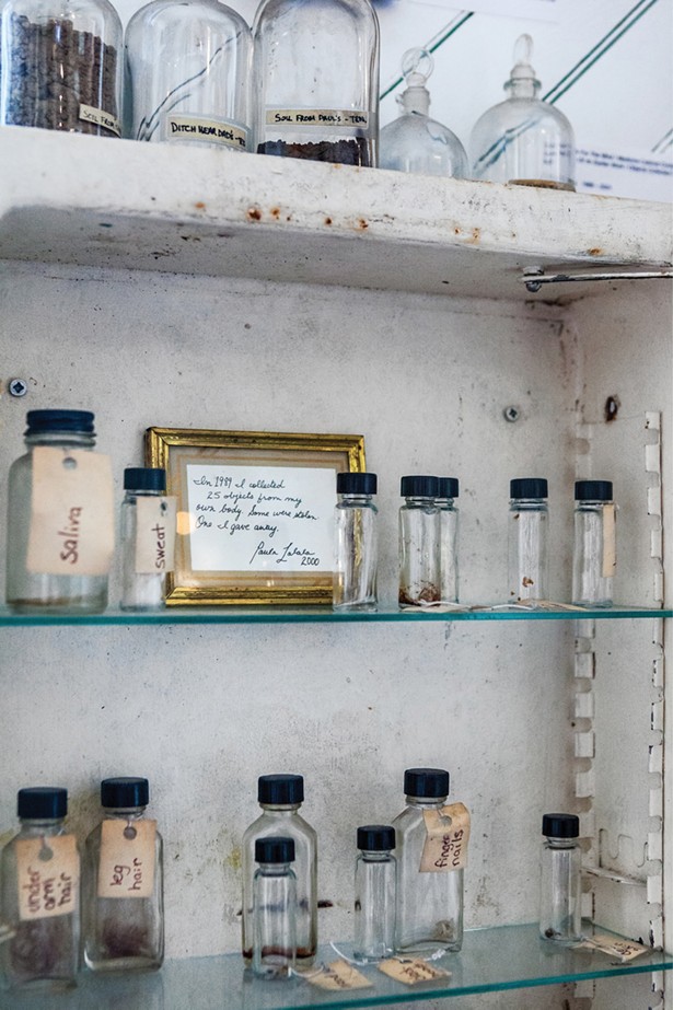 An antique metal medicine cabinet with a mirror labeled “touch me.” The framed message on the top shelf reads: “In 1989, I collected 25 objects from my own body. Some were stolen, one I gave away.” There are also vials containing water and dirt from her parents’ and siblings’ homes. - WINONA BARTON-BALLENTINE