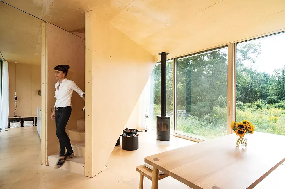 Betts descending the staircase from the second floor master suite into the home’s formal dining area. The couple finished all the home’s interior surfaces, including the built-in dining table, benches and kitchen island, with simple plywood. “We didn’t want anything to distract from the geometry of the space or the view of the landscape,” she explains. “But the plywood is beautiful in its own right—it has - its own configuration and pattern to the screws and the paneling.” - PHOTO: DEBORAH DEGRAFFENREID