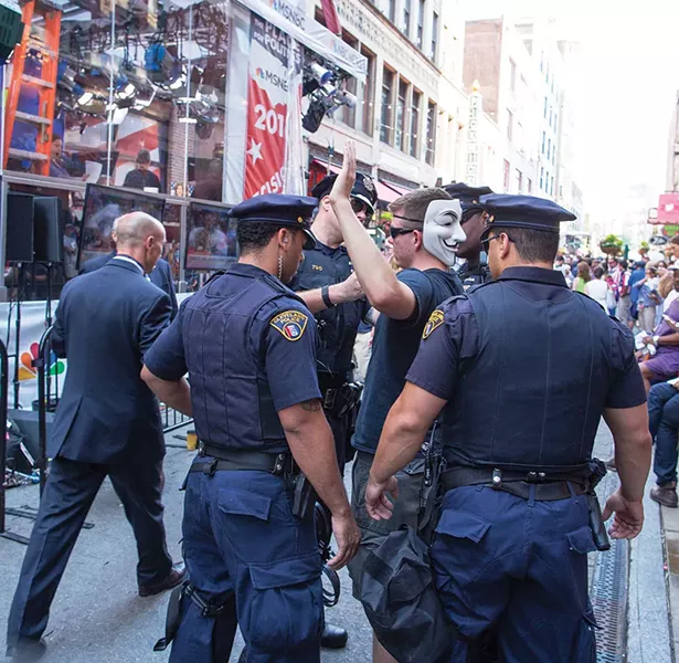 Police officers from across America (3500 in total) arrived in Cleveland to maintain order. Police questioned this bystander who was carrying a handheld electronic device that looked as if it could trip on an improvised explosive device (IED). The Guy Fawkes mask made him a profile target. - FRANK SPINELLI