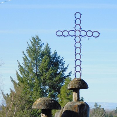 Two wooden mushrooms are carved out of two different tree stumps with a cross made of iron rings perched on top of the sculpture in a residential area of Genesee, Idaho on 3-19-24
