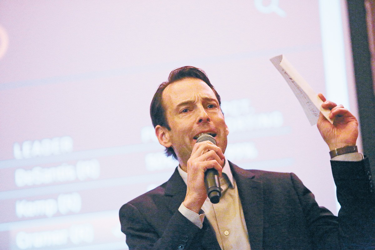 Against the blurred background of a projector screen, a man in the bottom of the frame speaks into a microphone while waving a piece of paper in the air. 