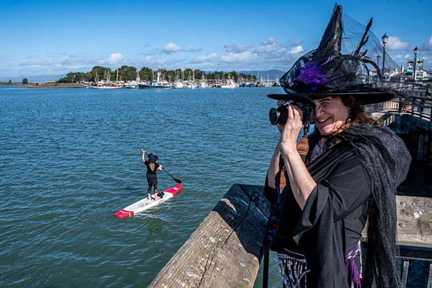 Dancing witch Lisa Wilhelmi Perkins, who chose to stay on land, photographed Ellen McLaughlin, of McKinleyville, on her red-and-white stand-up paddle board and other witch paddlers along the edge of Humboldt Bay with Woodley Island in the background.
