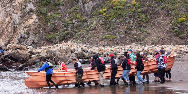 The nigilax̂ vessel is carried to the water by Native and non-Native community members at Sandy Cove in Fort Ross on May 27.