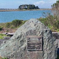 This National Historic Landmark plaque on Woodley Island, with Tuluwat Island (formerly Indian Island) in the background, merely refers to "Indian/Gunther Island, Site 67." No mention is made of the 1860 massacre.