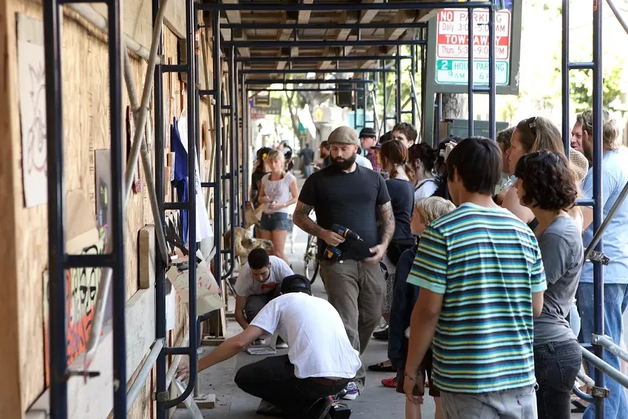 GUERRILLA ART! : Co-organizer Neal Breton of San Luis Art Supply (he’s the guy with the screw gun) helps donating artists mount their work on the wall of a construction barrier on Higuera Street, the scene of the last impromptu art show in which passersby were invited to take the work home for free. - PHOTO BY STEVE E. MILLER