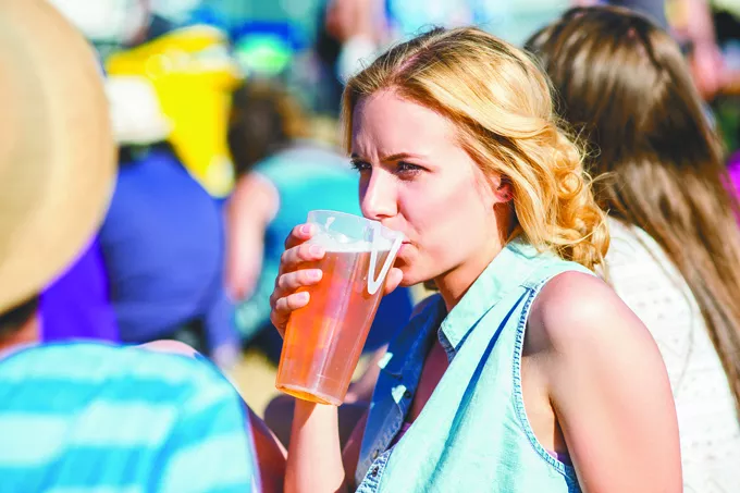 Young blond woman at summer music festival, drinking beer, sitting on the grass, crowded place - BIGSTOCK