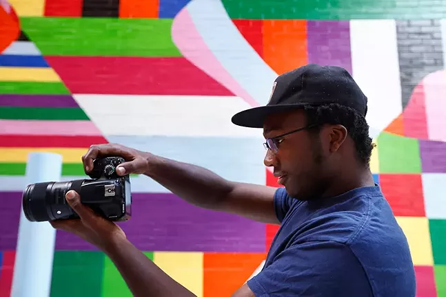 A profile of a man holding a camera to take a photo in front of a brightly colored painted mural