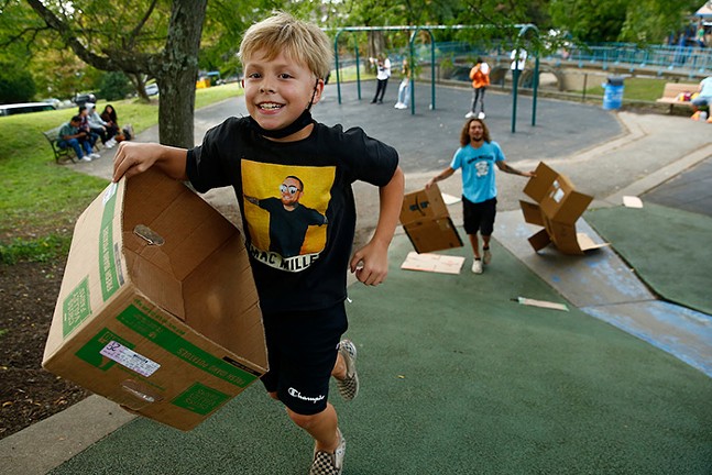 A young kid with a mask pushed down below his chin smiles as he carries a cardboard box up a hill in front of the blue slide he's going to ride down. An adult stands at the bottom of the blue slide holding two pieces of cardboard.