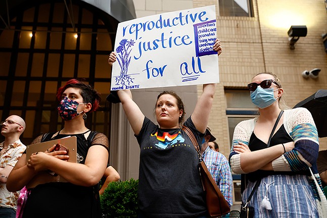 A woman holds up a sign that reads, - "reproductive justice for all" in between two other people wearing masks with their arms crossed