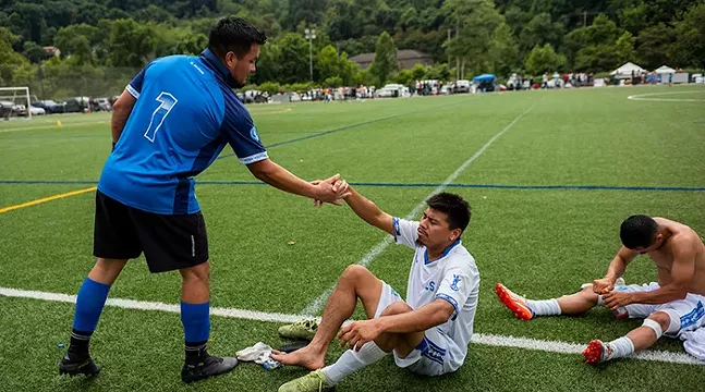 A soccer player reaches down to lend his hand to help another player sitting on the green playing field