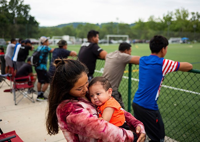 A woman holds a baby in her arms in front of a row of men leaning against a fence watching a soccer game