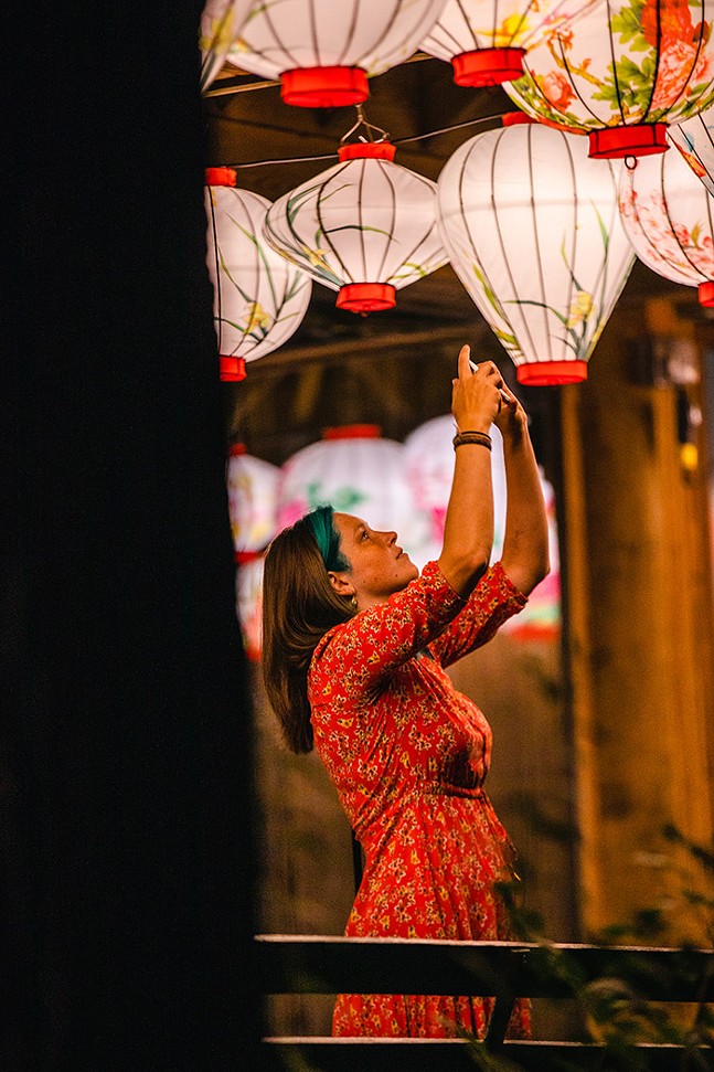 A woman raises her hands in the air to take a close-up cell phone photo of Asian lanterns hanging overhead