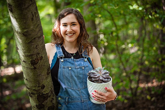 A white woman holds a potted plant and poses next to a tree.