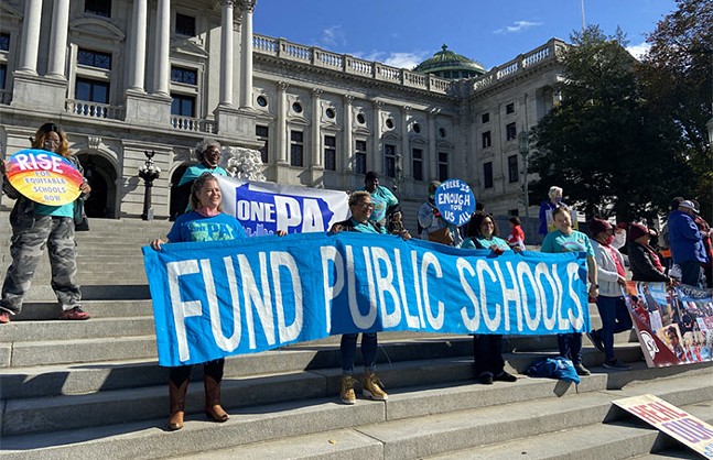A group of people stand on building steps holding signs including a large one held up by four people that says "Fund Public Schools"