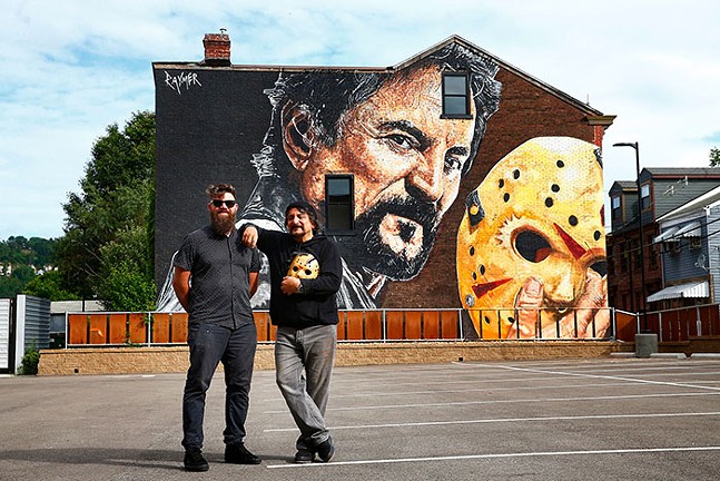 Two men stand side by side in front of a mural painted on the side of the building. One of the men in the foreground is painted holding a hockey mask on the mural