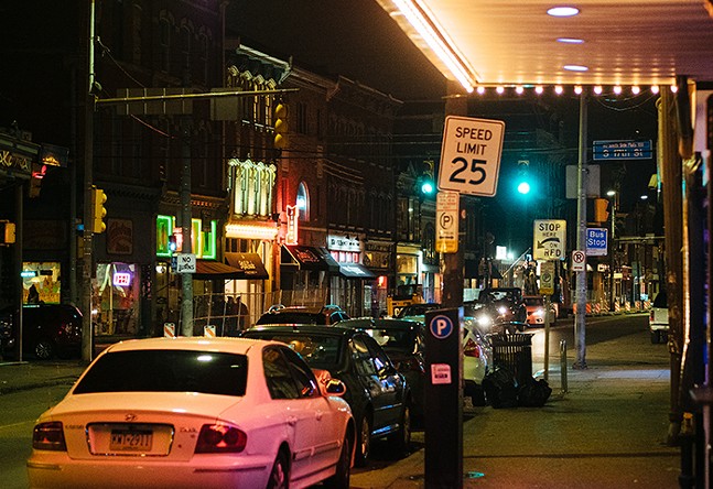 A photograph taken near the corner of South 17th Street and East Carson Street at night. Cars are parked on the right side of the street and businesses display neon signs
