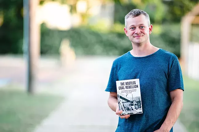 A smiling man in a blue shirt stands outside holding a copy of a book with "The Bootleg Coal Rebellion" written on the cover