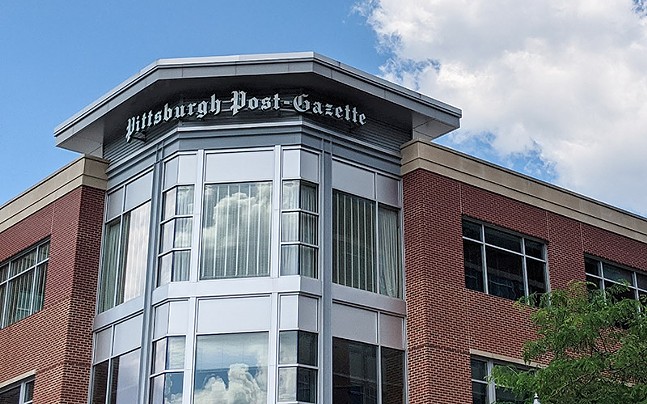 A red brick building with a metallic corner featuring a sign that says "Pittsburgh Post Gazette" sits under a blue sky.