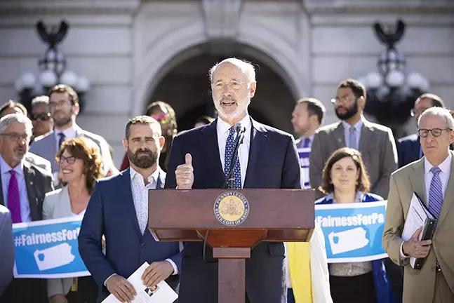 A man in a suit stands talking at a podium with a group of people behind him