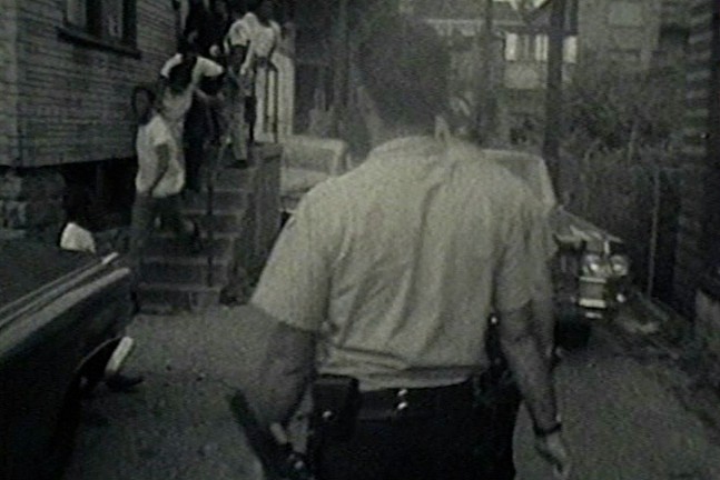 A blurry, black and white still from the documentary Pittsburgh Police 1969 shows a police officer looking on as a group of people exit a home.