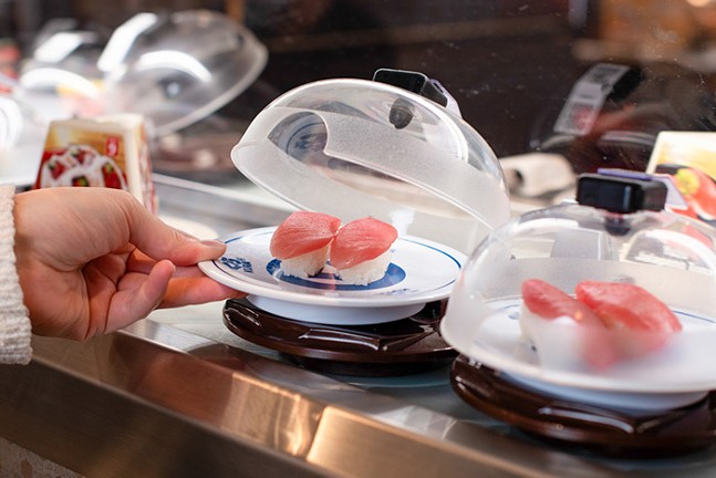 A hand reaches for a domed dish of sushi at Kura Revolving Sushi Bar.