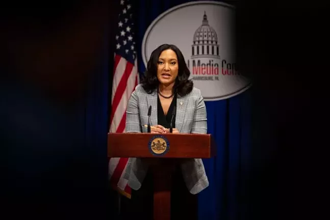 Woman standing and speaking at a podium in front of a blue curtain and an American flag