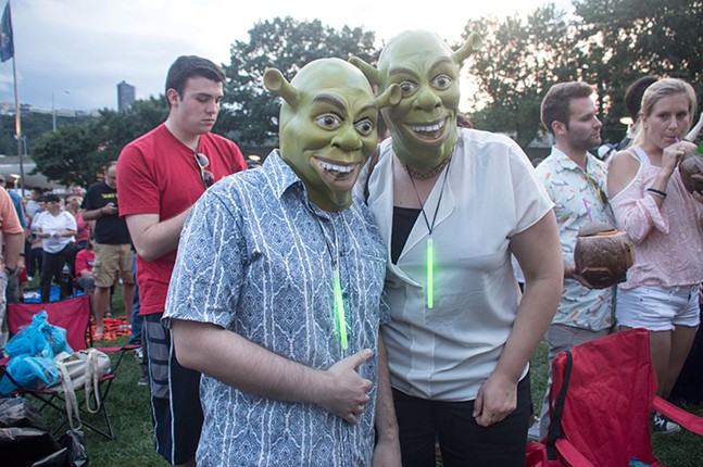 Two concert attendees wear Shrek masks and glo-stick necklaces, with one giving the thumbs up.
