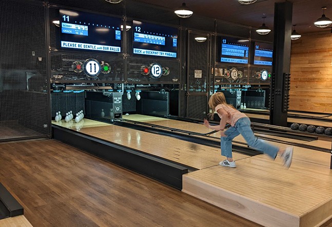 A young girl in a pink shirt and jeans throws a ball down a duckpin bowling lane at Pins Mechanical.