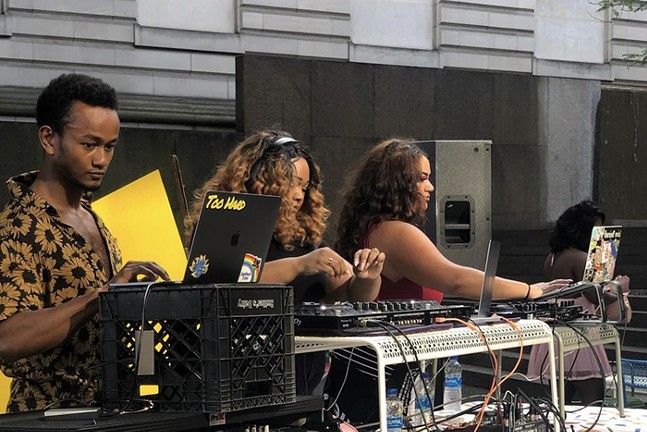 Three black DJs stand in a row behind a table with laptops, turntables and other equipment.