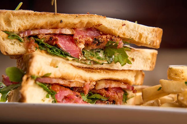 A sandwich with pink tuna, green arugula, and bacon is cut in half and displayed on a plate.