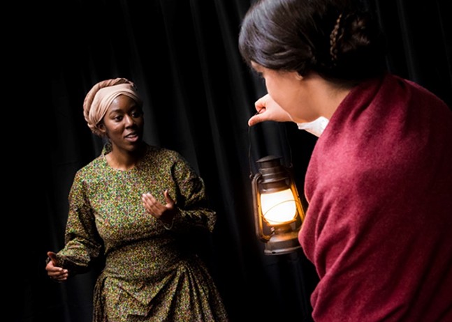 A Black woman dressed in period clothes portrays Harriet Tubman and another actor holds a lantern.