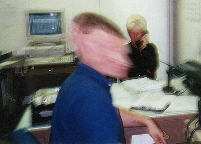 An extremely blurry images shoes two office workers, one looking towards the camera, another looking away as they talk on the phone.