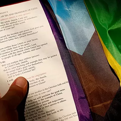 A hand holding the bible which has an LGBTQ flag draped over it