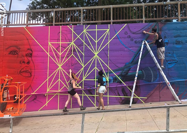 Artist Sarah Rutherford and youth apprentice Tehya Bollar tape off geometric lines while artist Justin Suarez applies paint to "This is Triumphant Music," the Wall\Therapy mural featuring Rochester musicians at Martin Luther King Jr. Park. - PHOTO BY REBECCA RAFFERTY