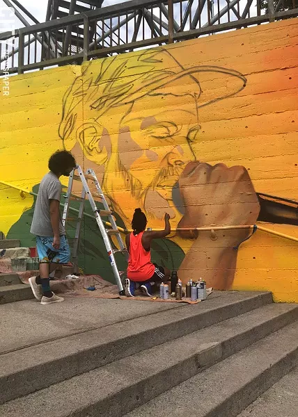 Youth apprentice Alex Baez Rivera looks on as artist Brittany Williams works on a section of the mural featuring Moses Rockwell. - PHOTO BY REBECCA RAFFERTY