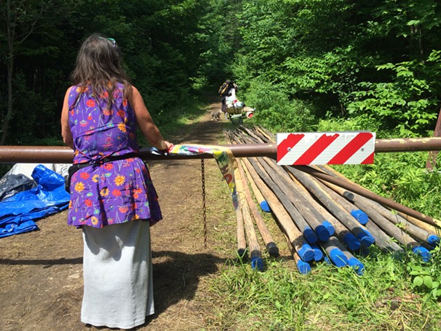 A woman who goes by “Feather” stands at the campsite entrance by poles that will be used to construct teepees. - MARK DAVIS