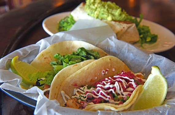 A carnitas taco is served gringo-style on a flour tortilla alongside a traditional fish taco on corn. Behind them is a pinto bean burrito with guacamole.