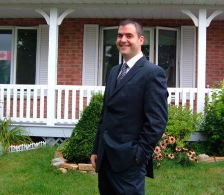 Aaron MacKenzie, a part-time teacher and full-time believer in trickle-up economics, stands in a suit in front of his house.